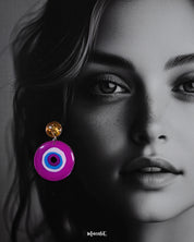 A close-up of a model wearing the Nigaahbaan stud earrings, showcasing the glittering gold studs and vibrant hand-painted evil eye design that pops against the skin—crafted to protect and impress in equal measure — Muselot.