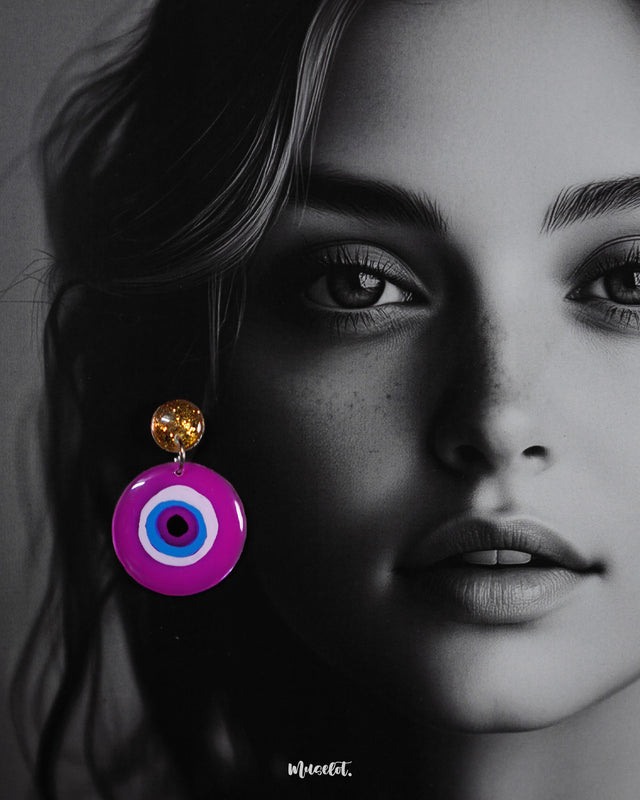 A close-up of a model wearing the Nigaahbaan stud earrings, showcasing the glittering gold studs and vibrant hand-painted evil eye design that pops against the skin—crafted to protect and impress in equal measure — Muselot.