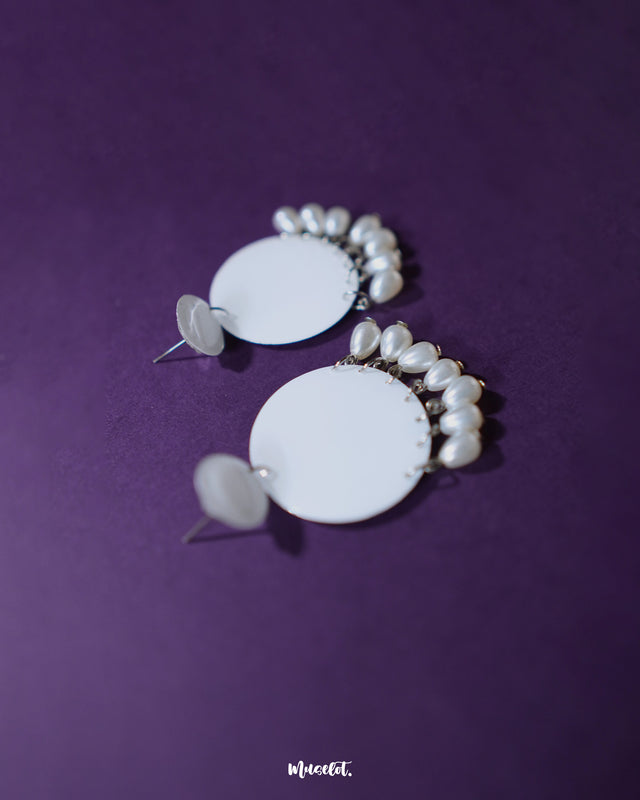 Noor white drop earrings displayed against a deep purple background, featuring glossy round tops with matching circular drops adorned with elegant pearl hangings, showcasing minimal elegance and highlighting timeless charm — Muselot.