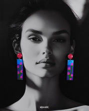 A woman wearing the Raatgul drop earrings brings them to life with effortless charm. The earrings’ glossy blue and purple panels, adorned with hand-painted pink buds and green stems, contrast beautifully with the bold red tops, adding a pop of artistic flair to her look — Muselot.