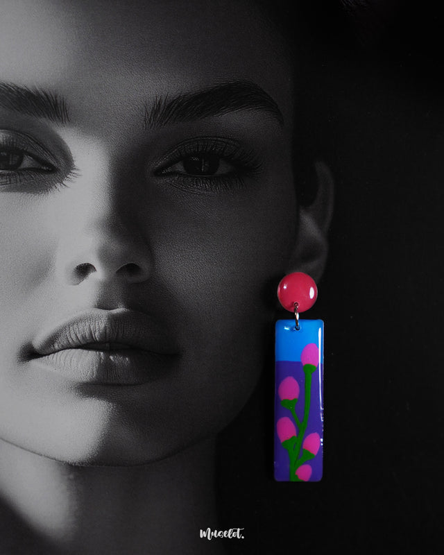 A close-up of a woman wearing the Raatgul drop earrings highlights their glossy, hand-painted design. The deep blue and violet panels feature delicate pink buds with green stems, anchored by a bold red stud, capturing the essence of handcrafted elegance — Muselot.