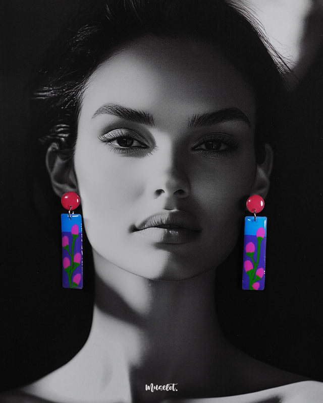 A woman wearing the Raatgul drop earrings brings them to life with effortless charm. The earrings’ glossy blue and purple panels, adorned with hand-painted pink buds and green stems, contrast beautifully with the bold red tops, adding a pop of artistic flair to her look — Muselot.