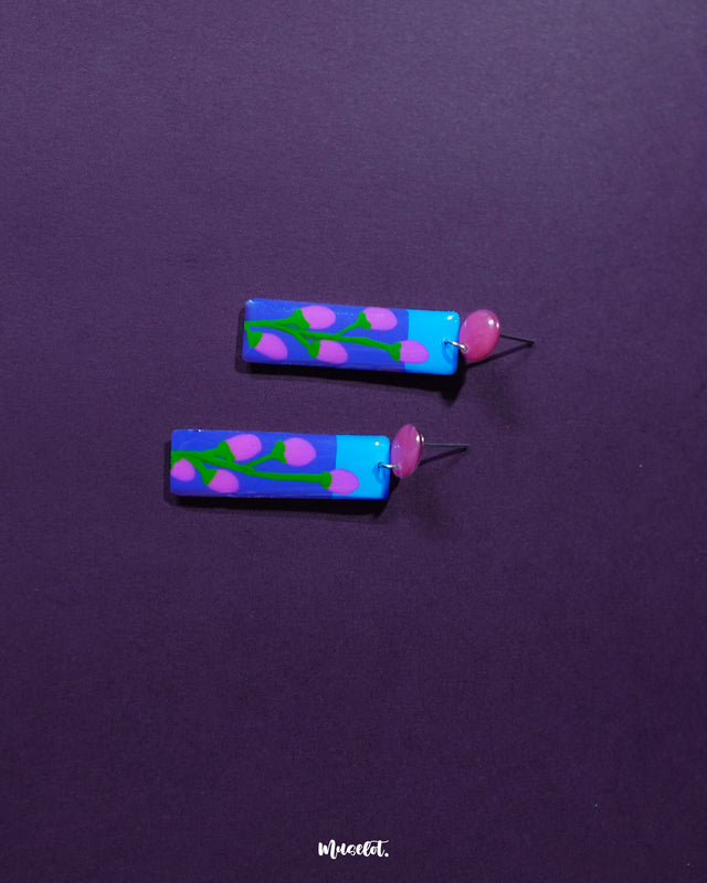 The Raatgul floral earrings rest against a rich purple backdrop, bringing out the vibrancy of their hand-painted details. With red studs and a striking blue-violet drop adorned with soft pink buds and green stems, these earrings evoke the charm of a midnight garden in bloom — Muselot.