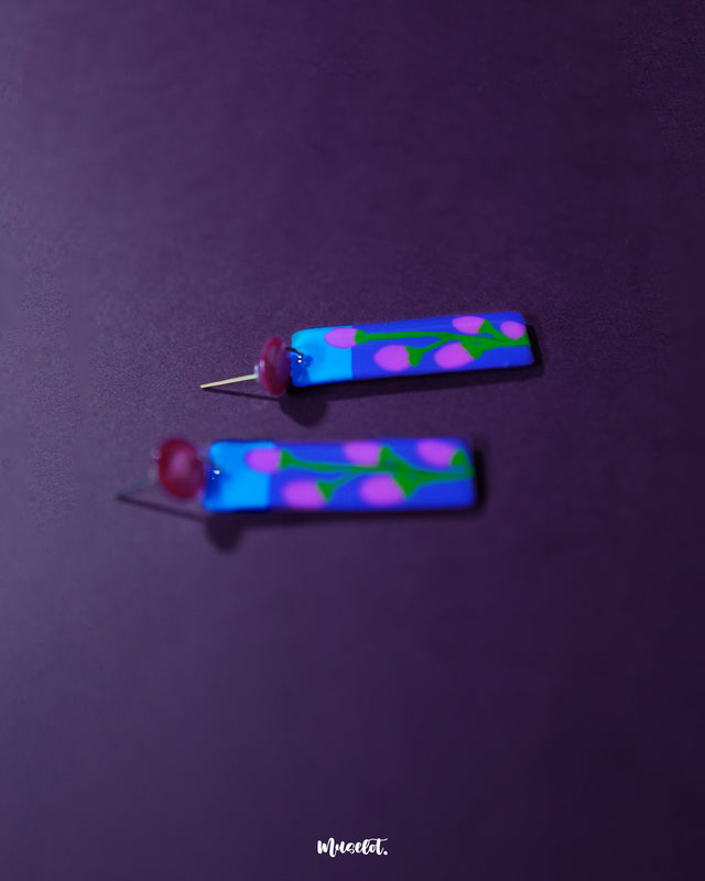 Close-up of Raatgul floral earrings showcasing the intricate hand-painted details—blush pink buds with green vines blossoming against a deep indigo backdrop, finished with glossy shine and bold red studs — Muselot.