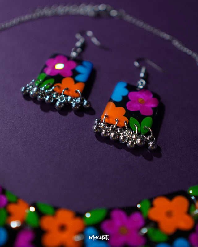 A close-up view of the Rangbaagh black floral earrings featuring delicate blue, orange, and purple flowers arranged in a balanced composition, paired with metallic gungroo drops that add a festive jingle and artisanal charm — Muselot