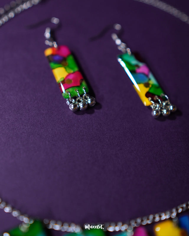 Colorful handcrafted jewelry set featuring a vibrant necklace and matching earrings with abstract multicolor patterns and silver bead accents, showcased on a deep purple background – Muselot.