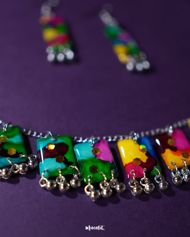 Close-up of the Rangbaari colorful jewelry set featuring vivid multicolor rectangular charms with silver bead accents on a silver chain necklace and matching earrings, highlighting the intricate patterns and vibrant hues – Muselot.