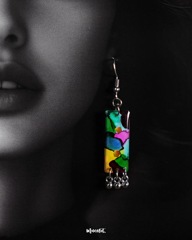 Close-up of a model wearing a vibrant rectangular earring from the Rangbaari colorful jewelry set by Muselot. The earring features a blend of bright green, pink, yellow, and blue hues, accented with small silver ghungroos at the bottom, standing out against the model’s dark hair and skin.