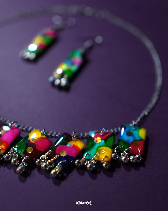 Rangbaari colorful jewelry set with a silver chain necklace and matching earrings, featuring bold multicolor abstract rectangular charms and delicate silver bead accents, artistically displayed on a deep purple background – Muselot.