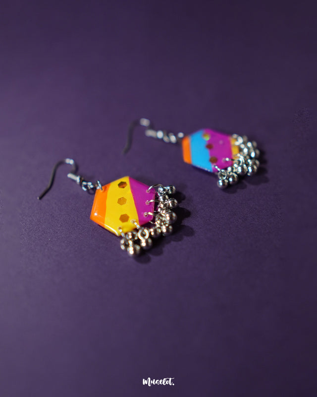 The Rangbahaar colorful hook earrings lie flat against a pastel surface, displaying their hexagonal form filled with vivid threadwork and shimmering sequins, finished with tiny silver bead danglers — Muselot.