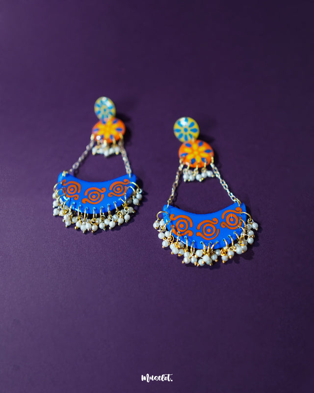 The Ranglahar blue chandbaali earrings rest against a rich purple background, enhancing their vibrant blue and orange enamel work. Golden chains and dainty pearl hangings add movement and charm, while the hand-painted floral studs bring a whimsical, folk-art-inspired elegance — Muselot.