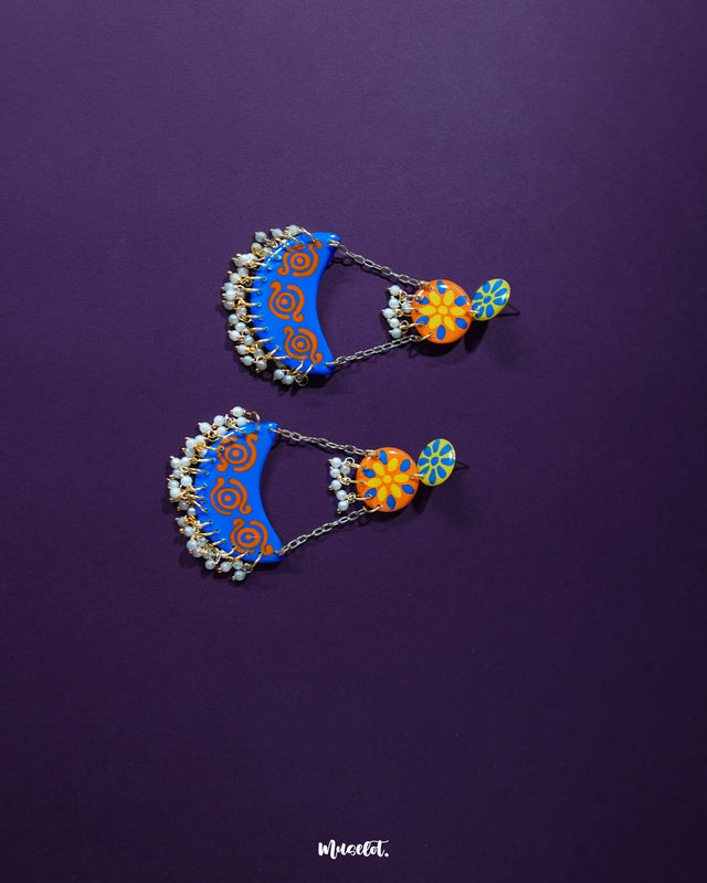 The Ranglahar blue chandbaali earrings showcase a playful harmony of vivid blue and orange hues, with hand-painted floral motifs and swirling patterns. Suspended by golden chains, the crescent design is adorned with delicate pearl drops that add a festive flair to these artistic statement pieces — Muselot.