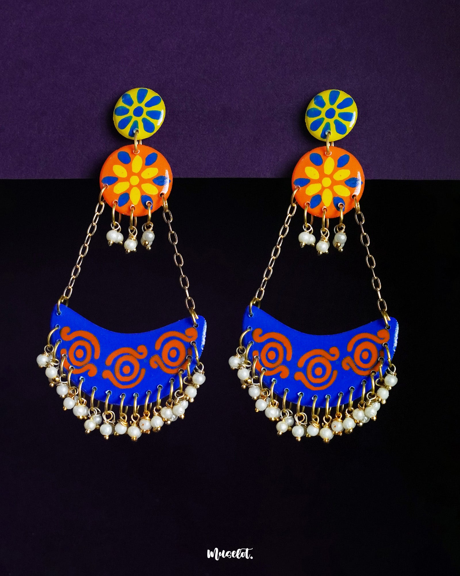 A vibrant pair of Ranglahar chandbali earrings featuring bold blue and orange crescent drops with intricate swirl patterns, suspended by golden chains from floral enamel tops, finished with tiny pearl danglers that add rhythmic charm and festive elegance — Muselot.