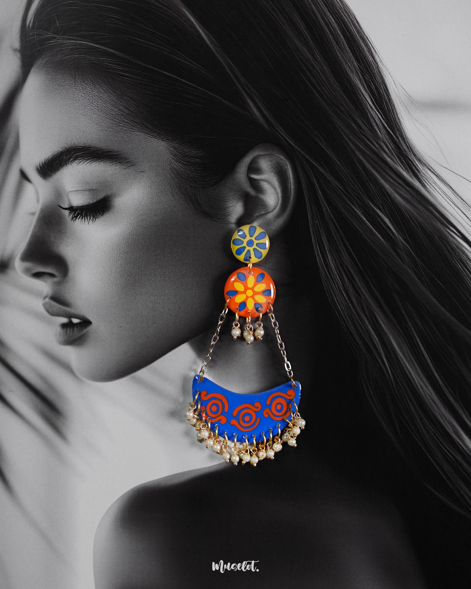 A woman wearing the Ranglahar chandbali earrings showcases their striking festive appeal—featuring bold blue and orange crescent drops adorned with intricate swirl motifs and delicate pearl hangings, all suspended from playful floral tops. The look blends traditional charm with a modern twist — Muselot.