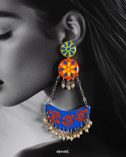 A close-up of a woman wearing the Ranglahar chandbali earrings highlights their vivid blue and orange enamel detailing, playful floral studs, and delicate pearl hangings. The intricate hand-painted patterns and gold-toned chains exude festive vibrancy with a contemporary edge — Muselot.