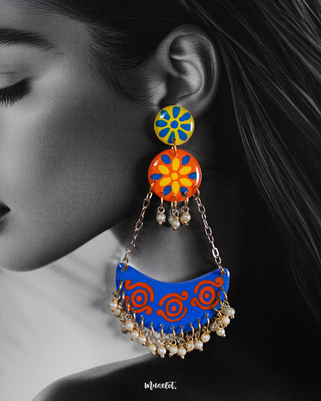 A close-up of a woman wearing the Ranglahar chandbali earrings highlights their vivid blue and orange enamel detailing, playful floral studs, and delicate pearl hangings. The intricate hand-painted patterns and gold-toned chains exude festive vibrancy with a contemporary edge — Muselot.