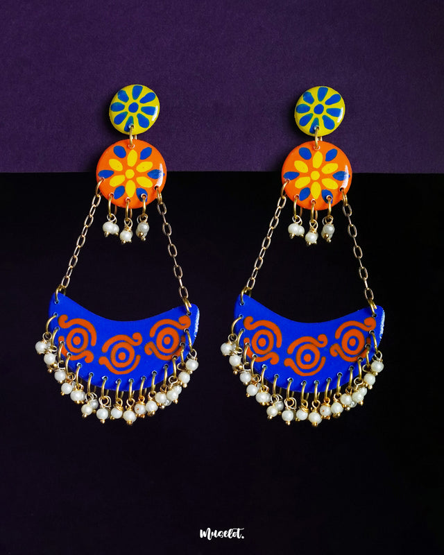 A vibrant pair of Ranglahar chandbali earrings featuring bold blue and orange crescent drops with intricate swirl patterns, suspended by golden chains from floral enamel tops, finished with tiny pearl danglers that add rhythmic charm and festive elegance — Muselot.