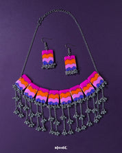 A vibrant handmade necklace and earring set featuring bold chevron patterns in pink, orange, purple, and blue, accented with silver-toned jhumki-style danglers — a playful fusion of tribal and modern aesthetics laid on a deep violet backdrop — Muselot.