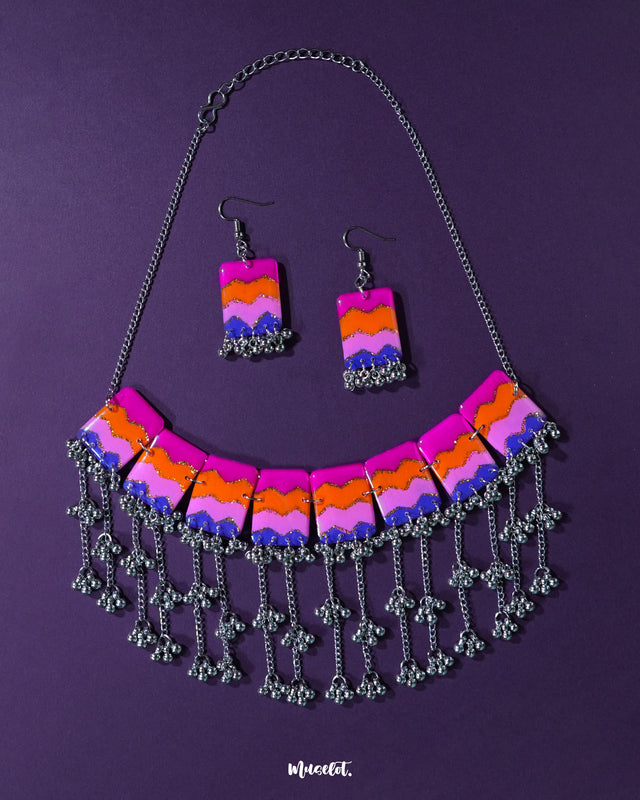 A vibrant handmade necklace and earring set featuring bold chevron patterns in pink, orange, purple, and blue, accented with silver-toned jhumki-style danglers — a playful fusion of tribal and modern aesthetics laid on a deep violet backdrop — Muselot.