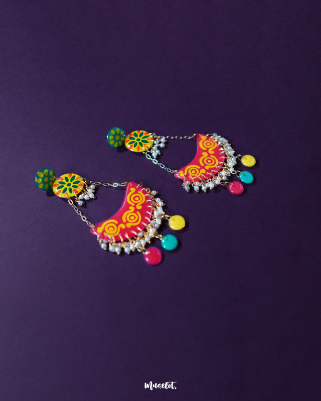 A pair of Rangsiya colorful chandbali earrings displayed against a deep purple background, highlighting their vivid hand-painted patterns, playful hanging beads, and traditional crescent design — Muselot.