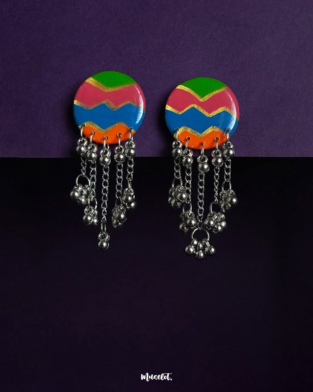 A vibrant pair of round handcrafted earrings featuring bold zigzag patterns in green, pink, blue, and orange, finished with dangling silver chains and clustered beads—bursting with festive charm and playful movement – Muselot.