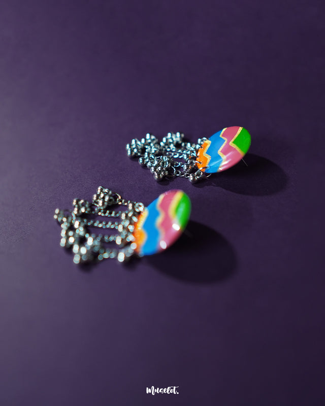 A pair of Rangtarang colorful long chain earrings displayed against a rich purple background, featuring bold zigzag patterns in green, pink, blue, and orange with dangling silver chains and clustered beads that bring a festive, boho vibe – Muselot.