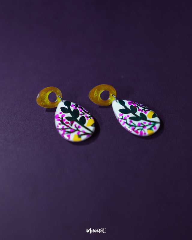 Raushanbahaar floral drop earrings resting on a deep purple background, showcasing teardrop-shaped dangles painted with pink buds, green leaves, and yellow accents, attached to shimmering golden open-loop studs — Muselot.