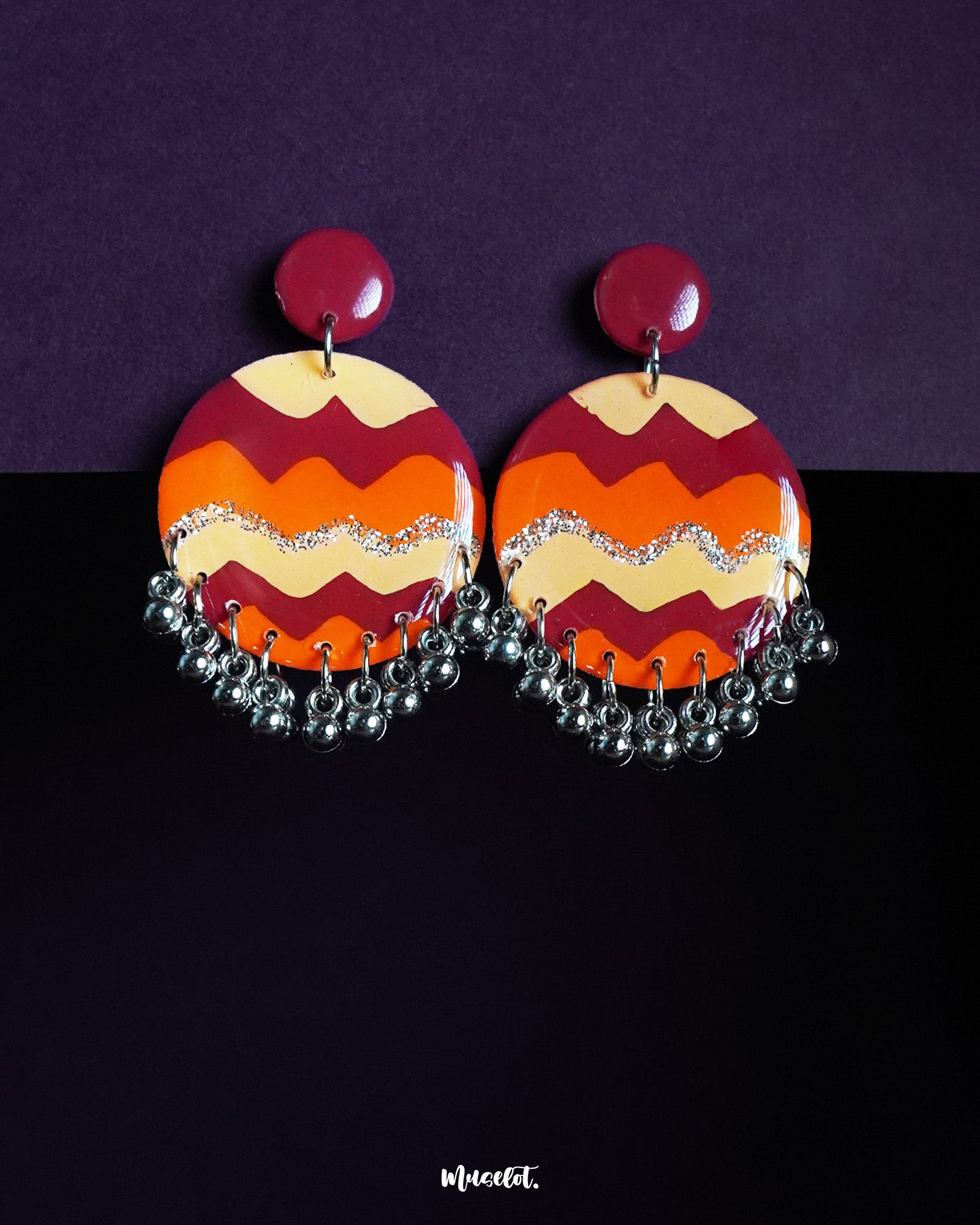 Sahar hook earrings featuring bold zigzag patterns in maroon, orange, and mustard with silver-tone ghungroos, set against a rich purple-black backdrop for a playful and festive charm — Muselot.