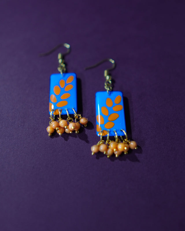 Sandhyaraag hook floral earrings placed on a purple background, featuring a bold blue base adorned with orange floral motifs and playful dangling beads that add a touch of boho flair — Muselot