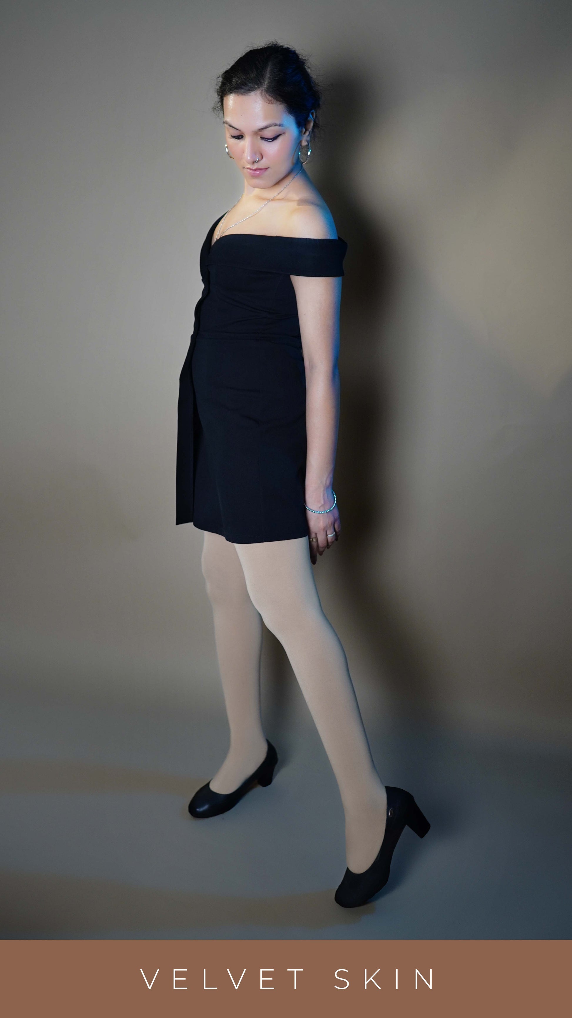 A woman wearing a stretchable semi opaque skin pantyhose, posing with her leg stretched at her side as she stands sideways as she looks at her tights, highlighting the trendy in vogue look-Muselot