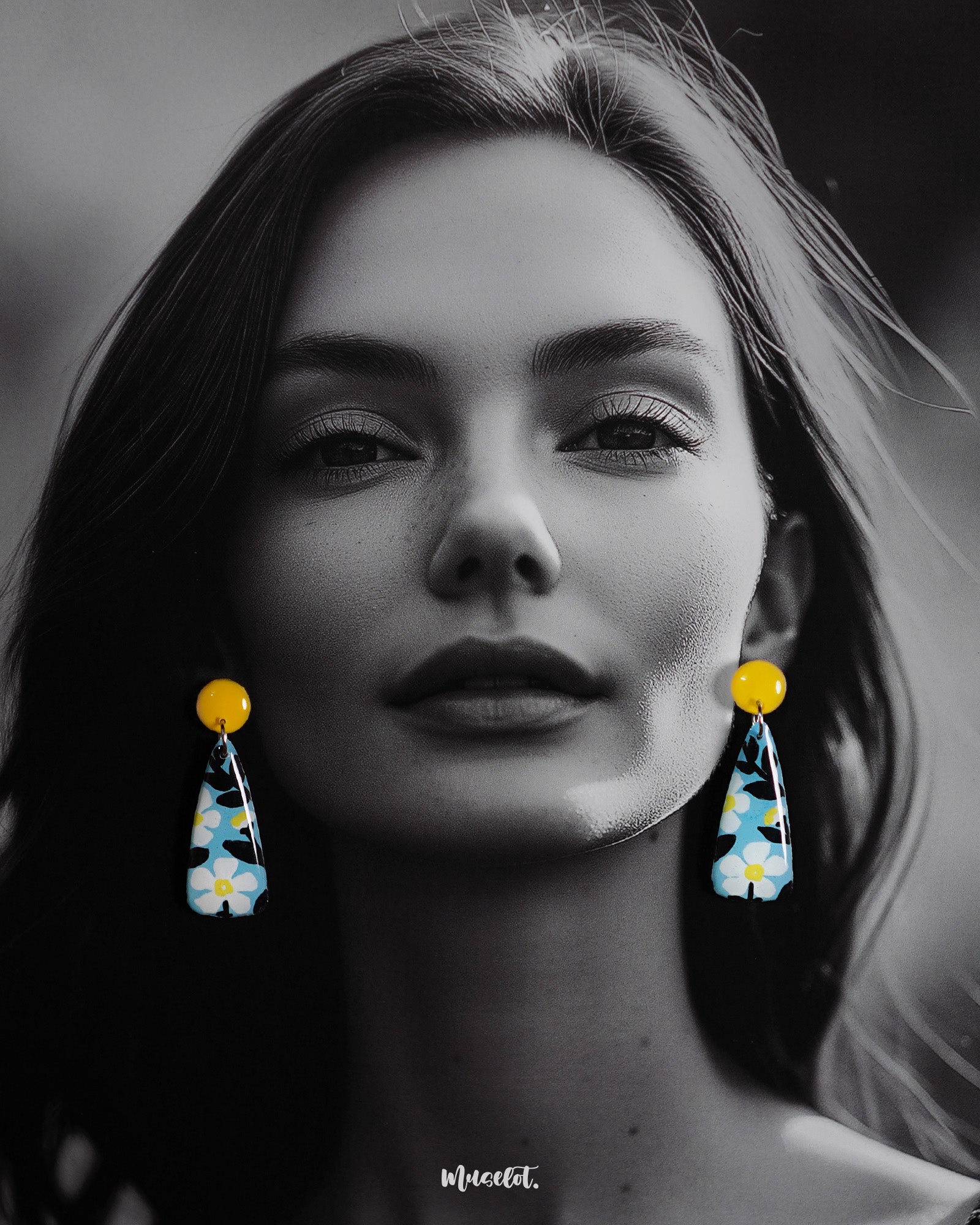 A model wearing the Shaamgul handmade drop earrings showcases their sleek, elongated shape adorned with hand-painted white flowers and black leaves on a pastel blue base, highlighting the bold pop of yellow from the round studs for a vibrant, artistic finish — Muselot.