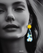 Close-up of a model wearing the Shaamgul handmade drop earrings, highlighting the smooth gloss finish and detailed floral artwork in white, black, and yellow tones on a soft blue base, showcasing the earrings’ hand-painted charm and bold yellow studs — Muselot.