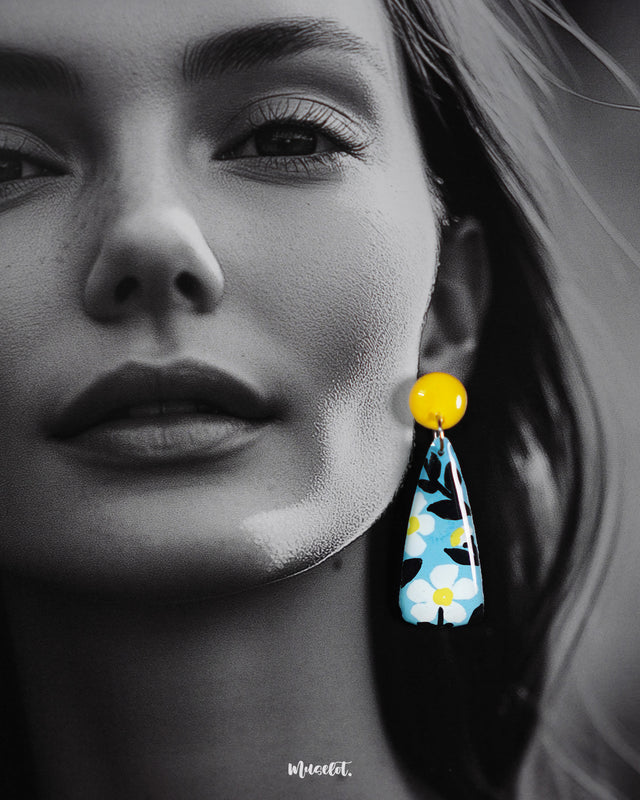 Close-up of a model wearing the Shaamgul handmade drop earrings, highlighting the smooth gloss finish and detailed floral artwork in white, black, and yellow tones on a soft blue base, showcasing the earrings’ hand-painted charm and bold yellow studs — Muselot.