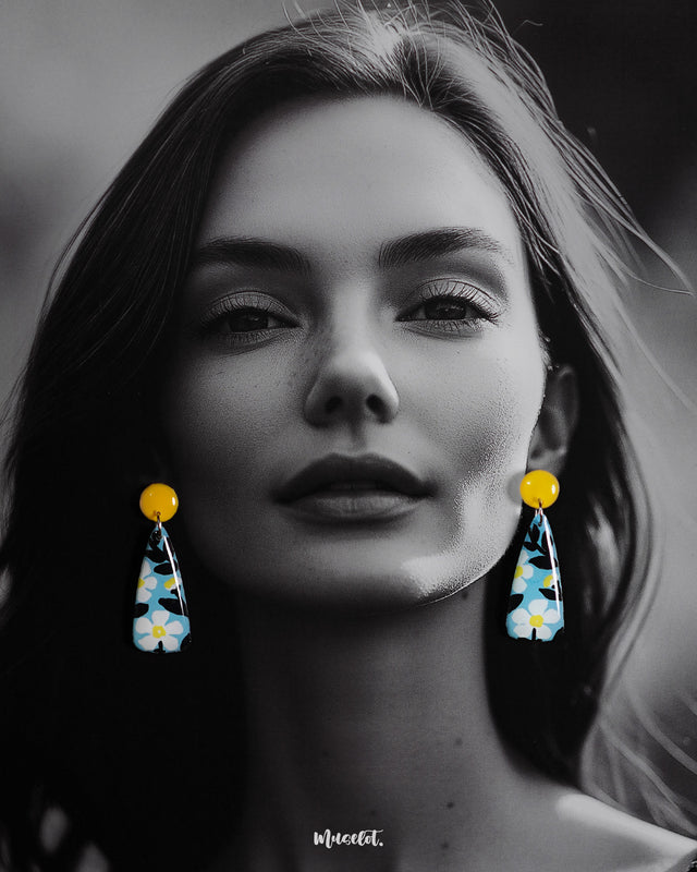 A model wearing the Shaamgul handmade drop earrings showcases their sleek, elongated shape adorned with hand-painted white flowers and black leaves on a pastel blue base, highlighting the bold pop of yellow from the round studs for a vibrant, artistic finish — Muselot.