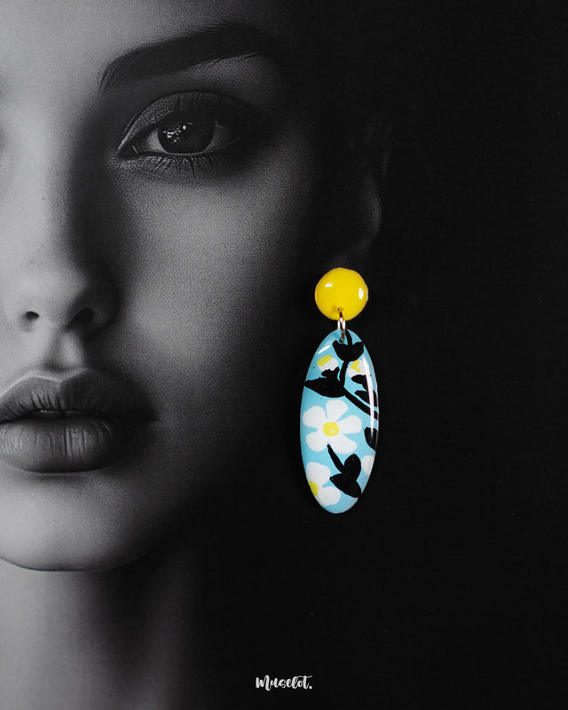 Close-up of a woman wearing the oval-shaped Shaamgul floral drop earrings, set against a rich purple background, showcasing the sky-blue base, hand-painted white daisies, black leafy details, and yellow stud for a striking, handmade statement — Muselot.