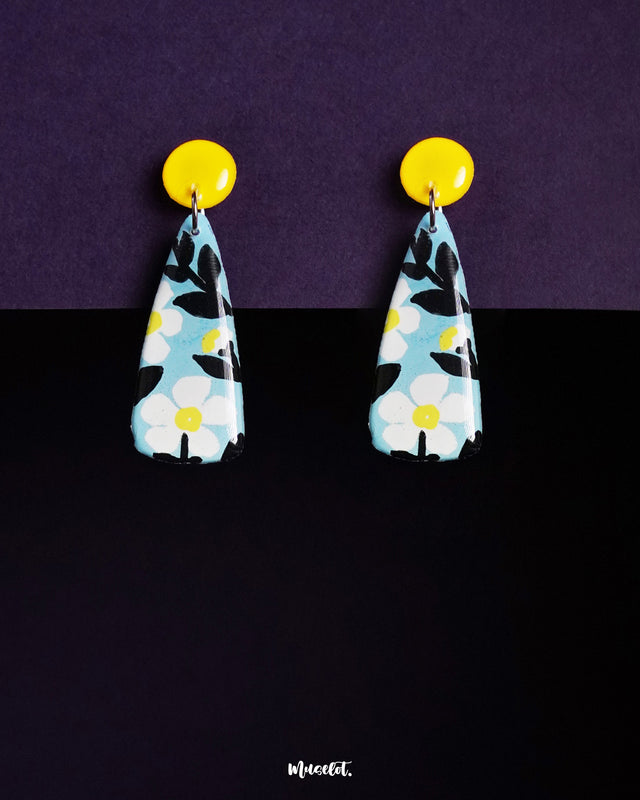 Shaamgul drop earrings featuring a tapered silhouette, hand-painted in soft blue with white floral motifs and bold black leaves, highlighting the artistic detail and showcasing the vibrant contrast with bright yellow round studs — Muselot.