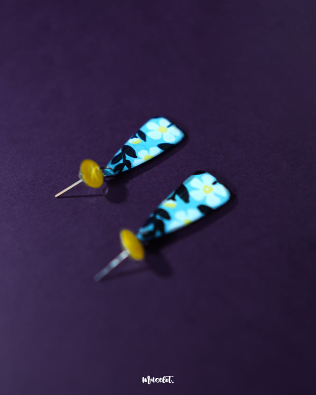 Shaamgul floral drop earrings in a soft blue hue, designed with delicate white flowers and black leaf accents, hanging from yellow studs — highlighting the intricate craftsmanship and cheerful charm of handmade accessories — Muselot.