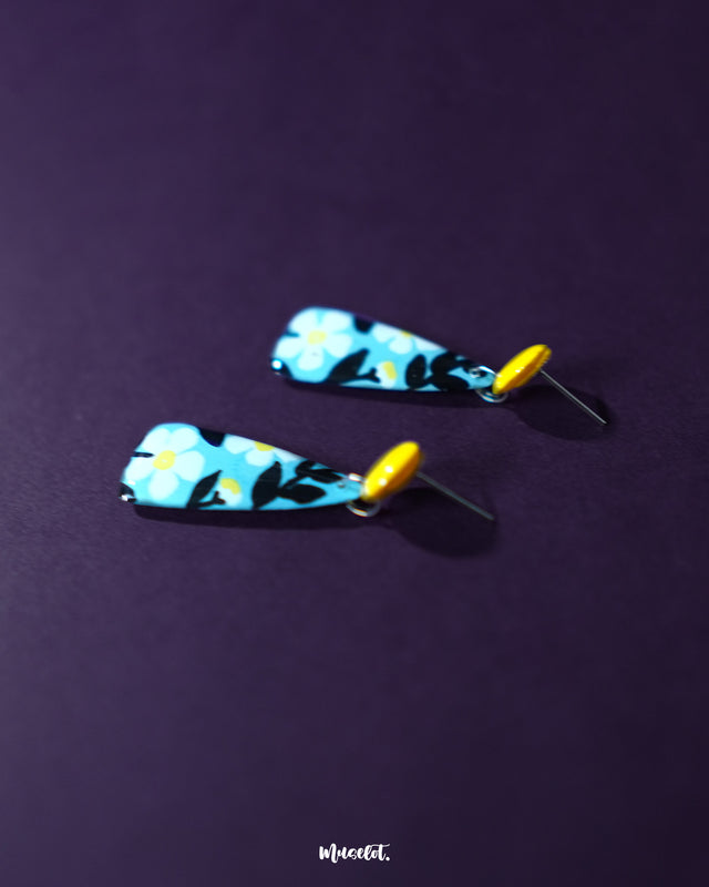 A pair of Shaamgul floral drop earrings featuring a blue teardrop base adorned with white and black floral accents, captured from a side angle to showcase their gentle curve and elegant fall — highlighting the graceful detailing of artisanal design — Muselot.