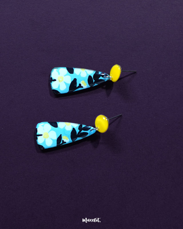 Shaamgul floral drop earrings laid flat on a soft surface, highlighting the intricate floral detailing in white and black atop the blue teardrop base,showcasing their handcrafted elegance and bohemian charm — Muselot.