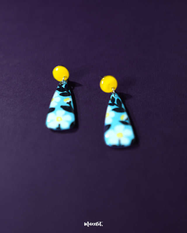  Shaamgul floral drop earrings placed against a purple background, highlighting the pastel blue base adorned with white daisies and black leaves, complete with a sunny yellow stud and fine detailing that showcases its handmade charm — Muselot.