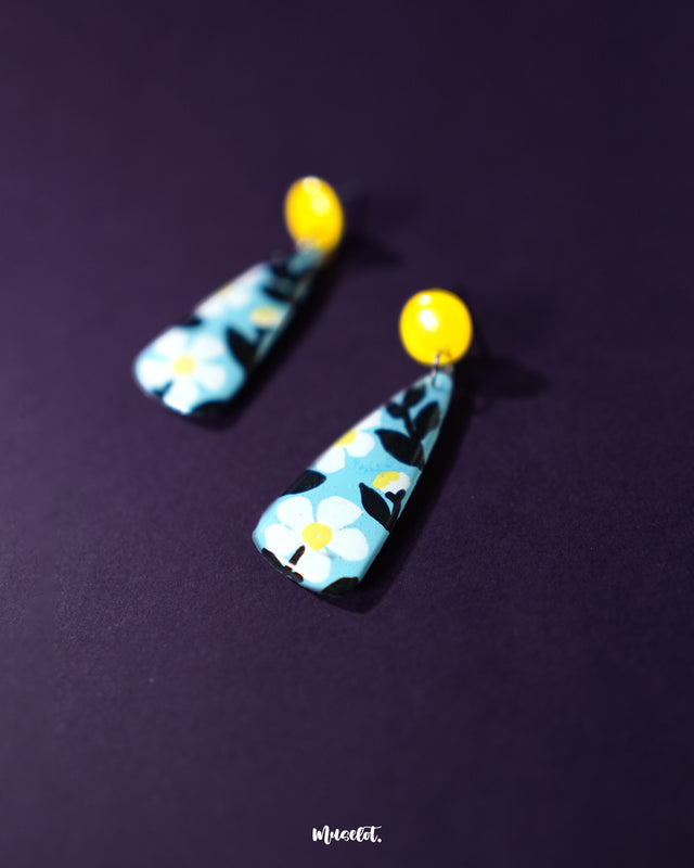 Shaamgul floral drop earrings resting on fabric, showcasing their unique shape with black and white floral accents and a soft blue drop, highlighting the handcrafted design — Muselot.