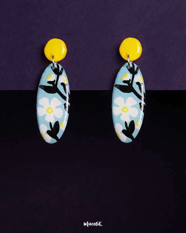 Oval-shaped Shaamgul floral drop earrings displayed against a rich purple background, featuring a sky-blue base with white daisy motifs, bold black leaves, and a striking yellow circular stud, creating a charming contrast — Muselot.