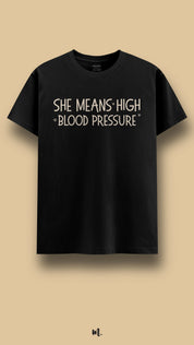 Black graphic t-shirt showcasing the statement “She Means High Blood Pressure” in clean beige typography with subtle heart accents, highlighting a sarcastic yet affectionate couple joke against a neutral beige background – Muselot