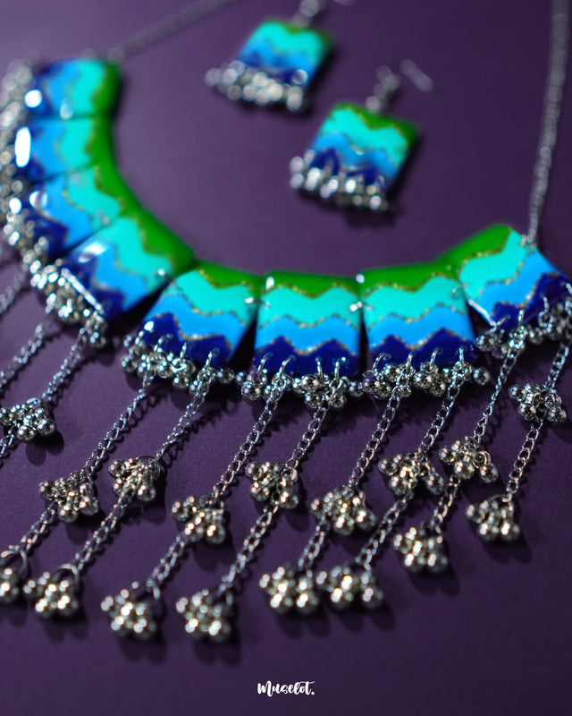Waterfall boho jewelry set placed on a deep purple background, highlighting its vibrant enamel panels in green, aqua, and navy with a touch of glitter. The silver chains and bead clusters showcase a flowing, layered design that gives a rhythmic and stylish look — Muselot.