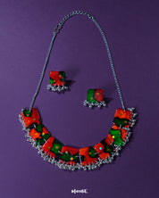 A vibrant handmade jewelry set,featuring a statement necklace and matching square earrings, crafted with bold red and green abstract patterns accented by tiny reflective sequins. The necklace is adorned with delicate metallic beads along the bottom edge, all set against a deep purple background — Muselot.