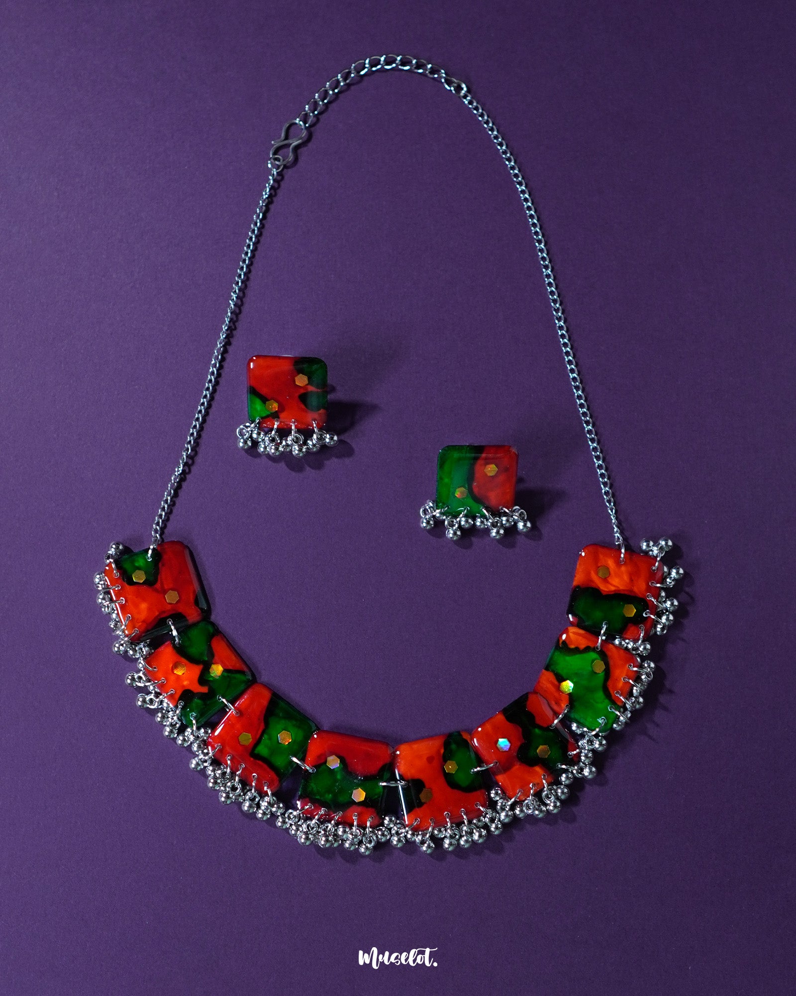 A vibrant handmade jewelry set,featuring a statement necklace and matching square earrings, crafted with bold red and green abstract patterns accented by tiny reflective sequins. The necklace is adorned with delicate metallic beads along the bottom edge, all set against a deep purple background — Muselot.