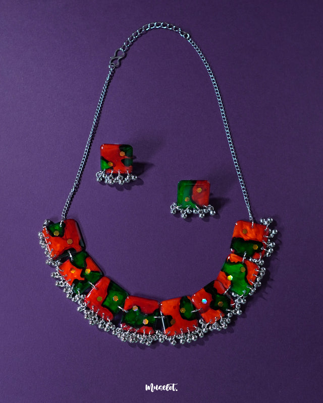 A vibrant handmade jewelry set,featuring a statement necklace and matching square earrings, crafted with bold red and green abstract patterns accented by tiny reflective sequins. The necklace is adorned with delicate metallic beads along the bottom edge, all set against a deep purple background — Muselot.