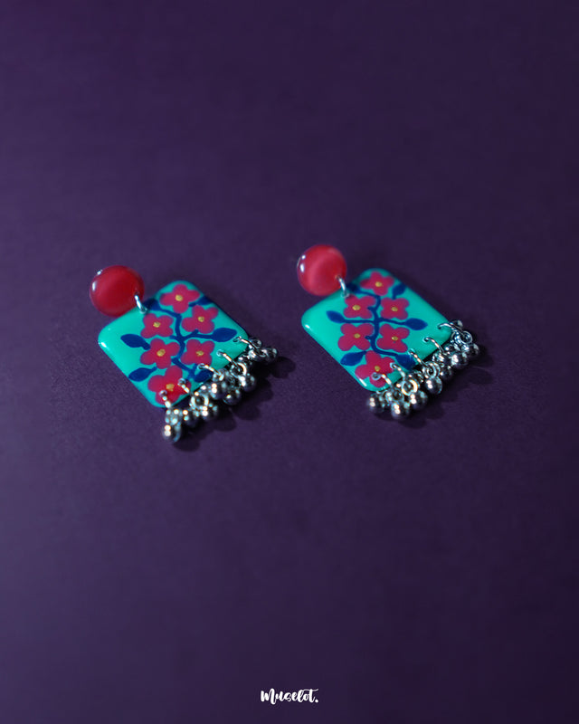 Surkhili floral earrings displayed against a purple background, showcasing the turquoise petals with red floral hand-painting and tiny silver ghungroos hanging below — Muselot.