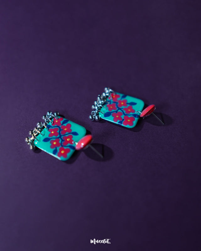 Pair of Surkhili floral earrings laid flat, highlighting the vibrant red blooms with delicately hand-painted petals and tiny dangling silver beads that add a charming ethnic flair — Muselot.
