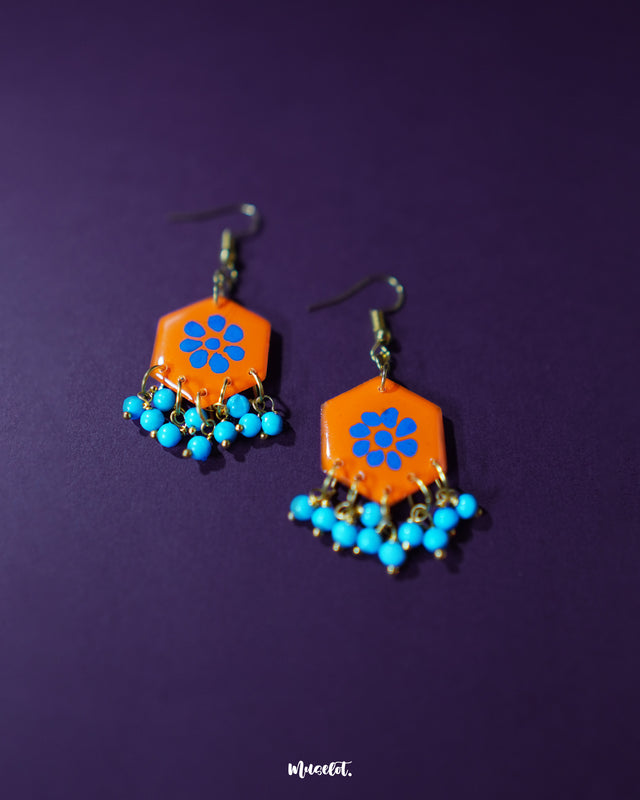 Surkhshaam orange floral stud drop earrings, displayed against a rich purple background, featuring vibrant blue floral accents and a graceful drop design that enhance their bold yet elegant charm — Muselot.